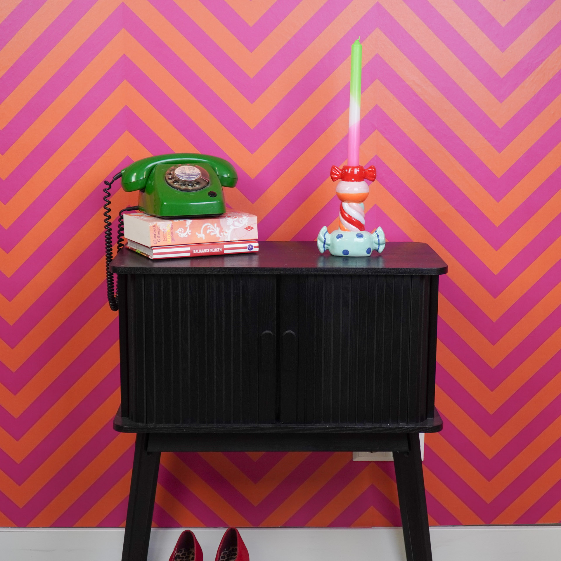 Aparador preto com telefone verde, livros e castiçal colorido contra parede de padrão zigzag cor-de-rosa e laranja