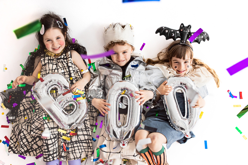 Três crianças com fantasias seguram balões metálicos com a palavra BOO rodeadas por confetes.