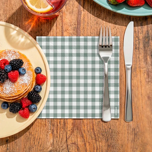 Guardanapo xadrez verde e branco com talheres metálicos sobre mesa de madeira, perto de prato com panquecas e frutos vermelhos.