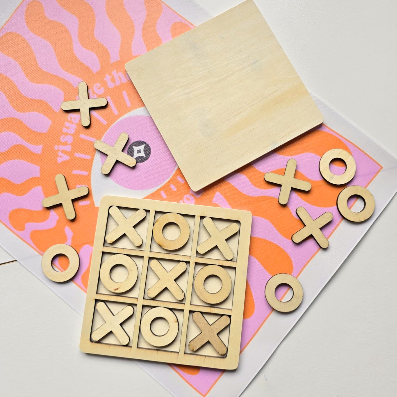 Jogo da velha em madeira clara com tabuleiro e peças X e O, fundo laranja e rosa com texto parcial.