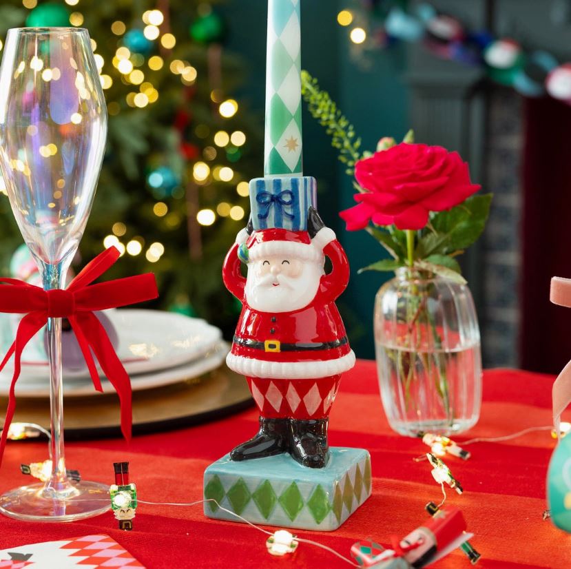Castiçal Pai Natal com vela em padrão de losangos, copo com laço, rosa vermelha em vaso e luzes decorativas numa mesa vermelha