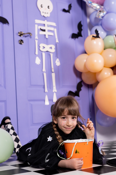 Criança com balde de Halloween num cenário decorado com caveira e balões.