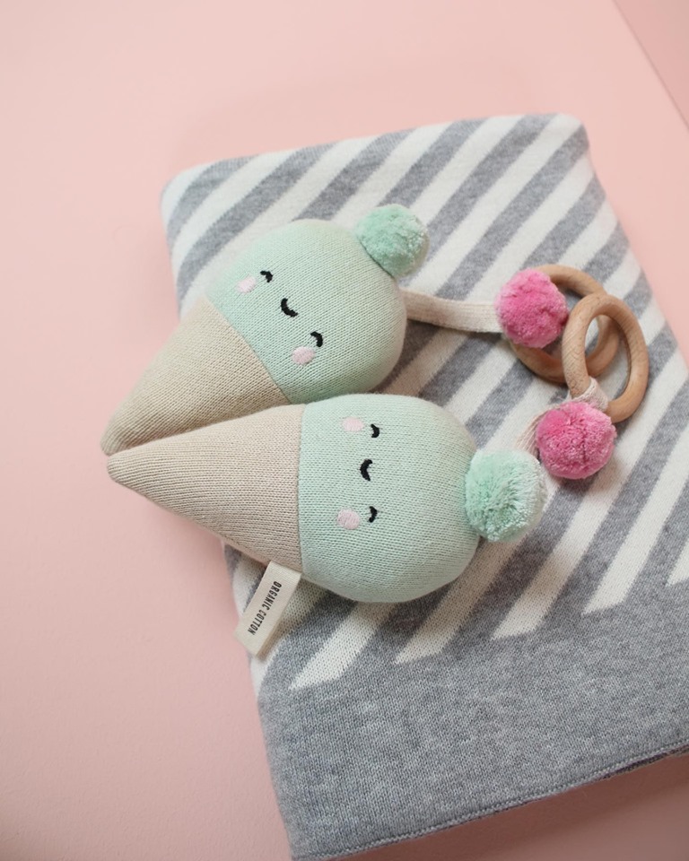 Brinquedo para bebé duplo com pompons e argolas de madeira sobre manta cinza às riscas em fundo rosa