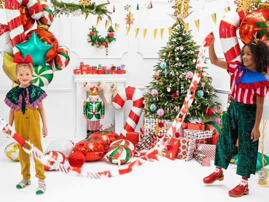Crianças com roupas coloridas brincam com varas de doces gigantes numa sala decorada para o Natal.