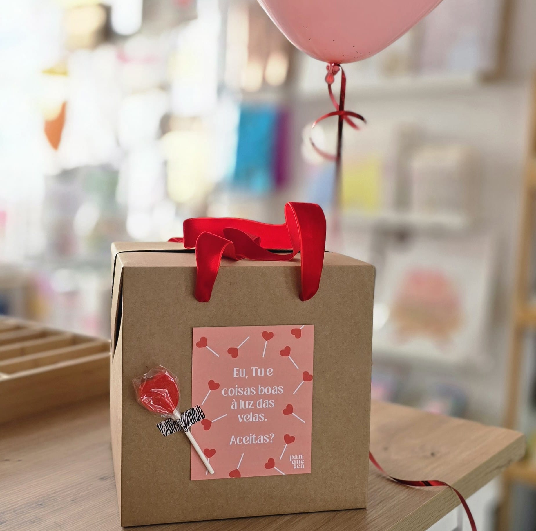 Dia dos Namorados - Não é uma caixa, é um convite delicioso! Caixa castanha com selo rosa e pirulito vermelho, em mesa de madeira, com bexiga cor-de-rosa