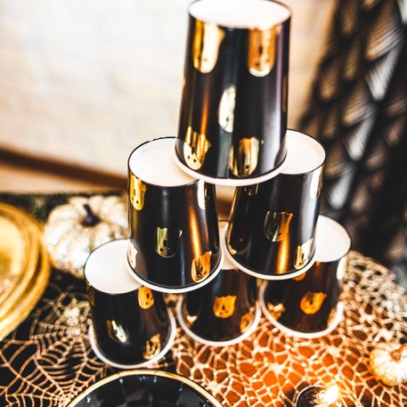 Seis copos pretos brilhantes empilhados em pirâmide numa mesa com toalha de teia de aranha e abóbora decorativa