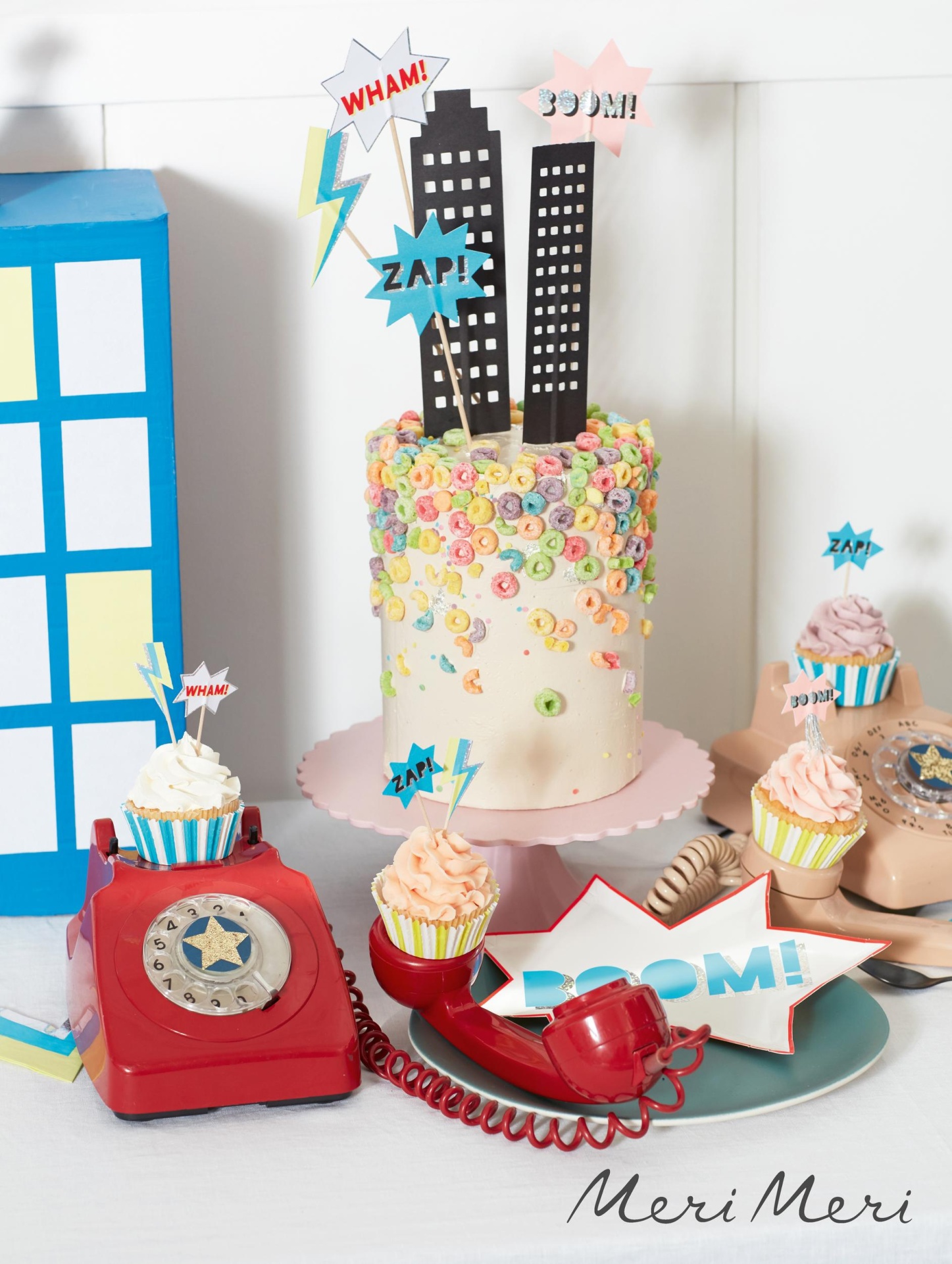 Bolo decorado com cereais coloridos e toppers de prédios e palavras em estilo de quadrinhos, com telefones antigos vermelhos e cupcakes ao redor