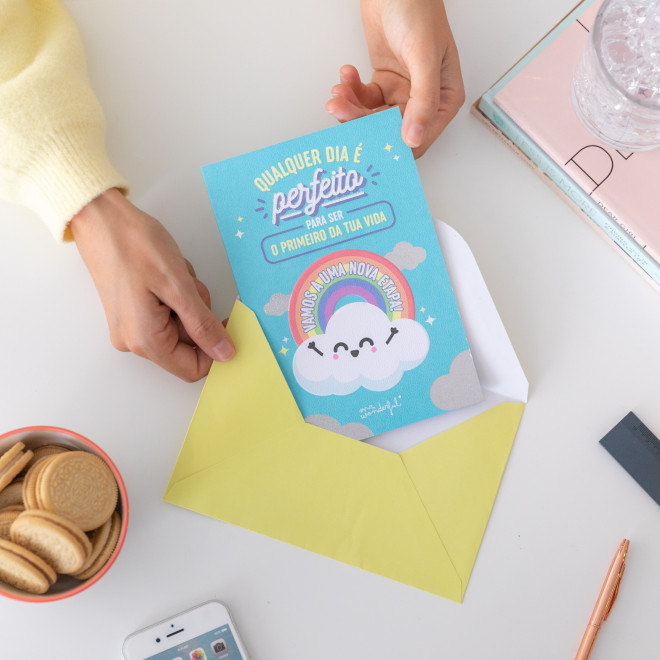 Cartão azul com nuvem e arco-íris dentro de envelope amarelo sobre mesa com biscoitos, telemóvel e copo