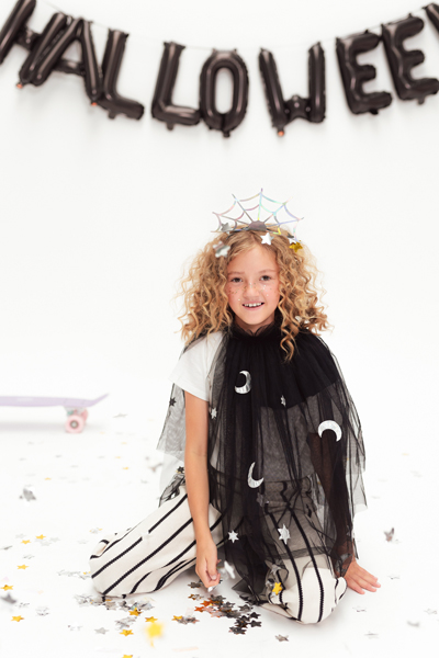 Menina com capa preta de Halloween e tiara com teia de aranha sentada em ambiente festivo com balões pretos
