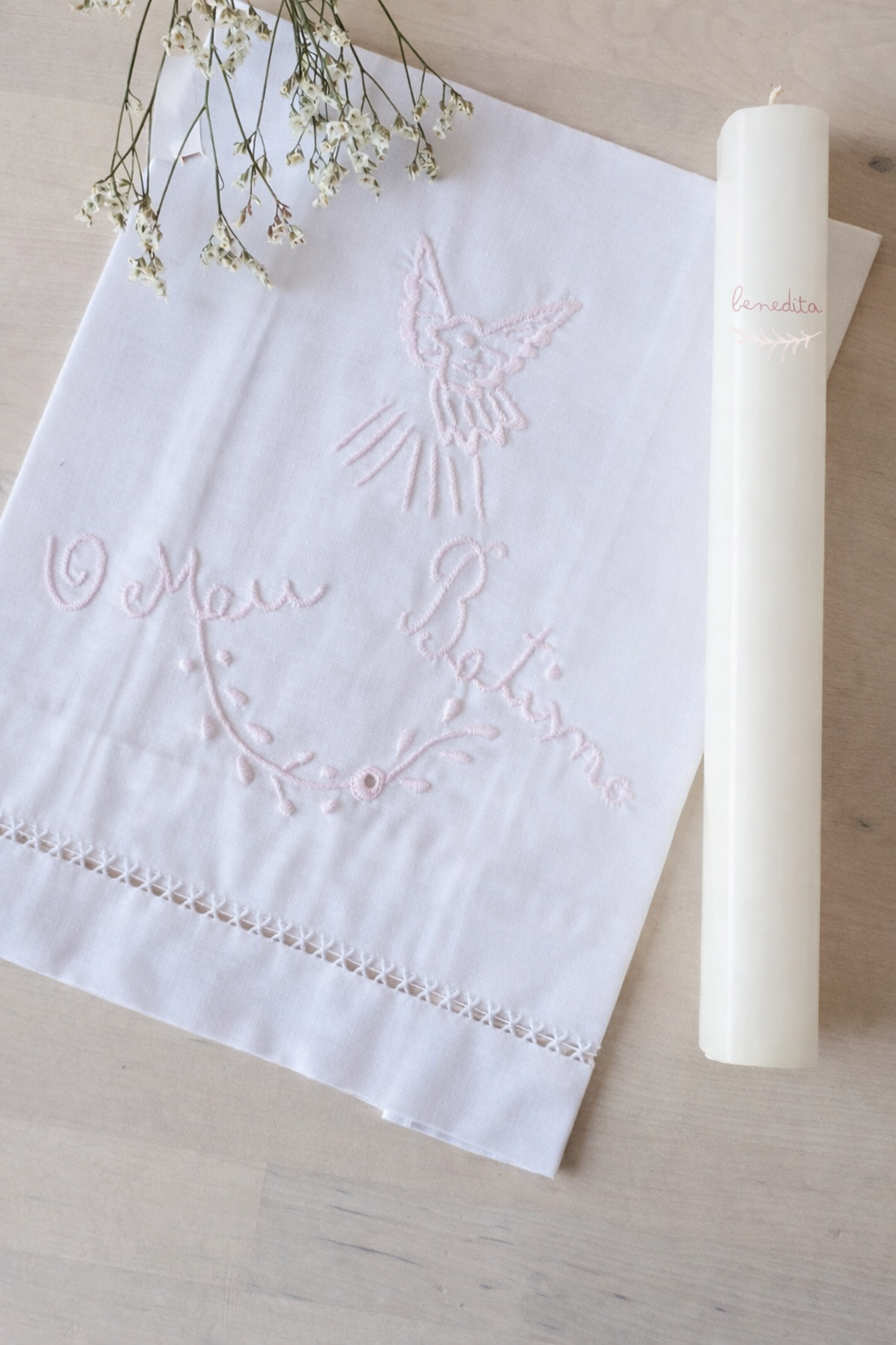 Toalha branca bordada com pomba e texto em rosa junto a uma vela branca com texto benedita