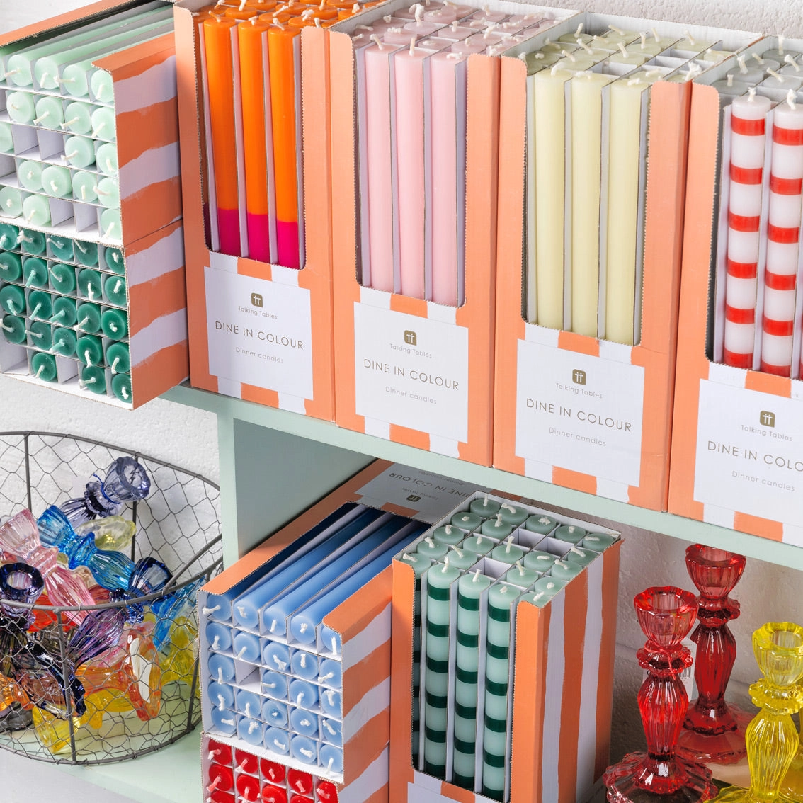 Velas de jantar variadas em caixas laranja branco com etiquetas, numa prateleira com castiçais coloridos ao lado.