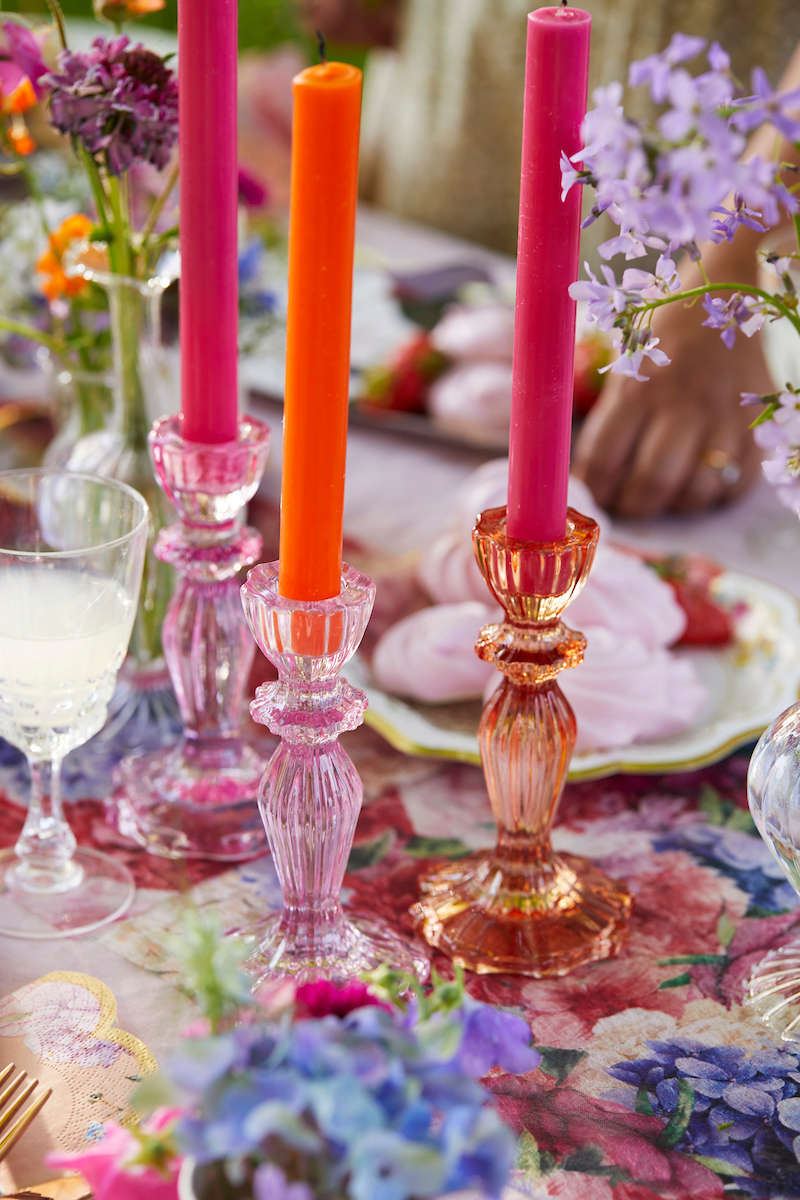 Castiçais de vidro rosa com velas coloridas numa mesa com toalha floral e flores