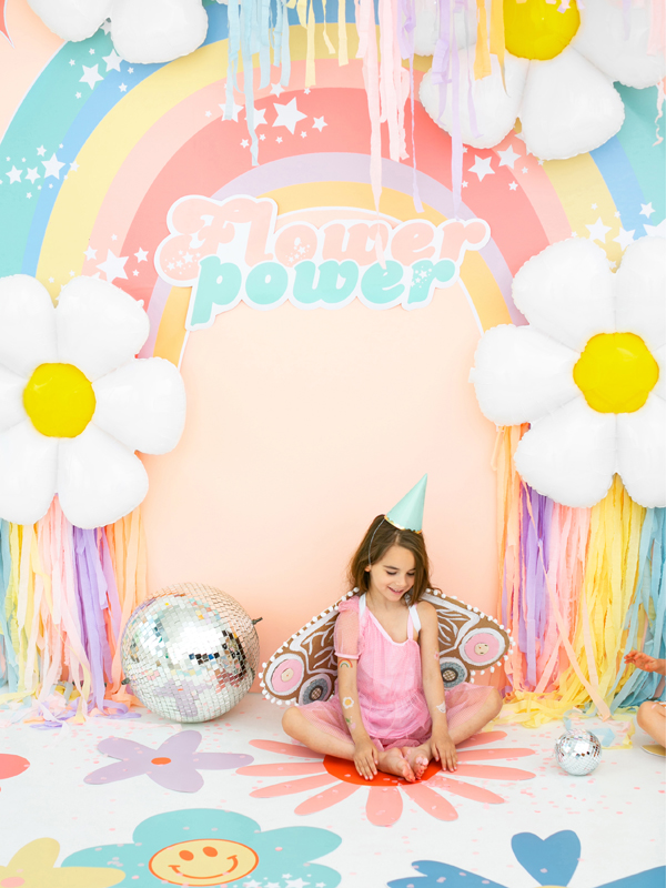 Decoração com tema Flower Power, arco-íris, flores grandes e menina em fantasia cor-de-rosa sentada no chão com decoração colorida