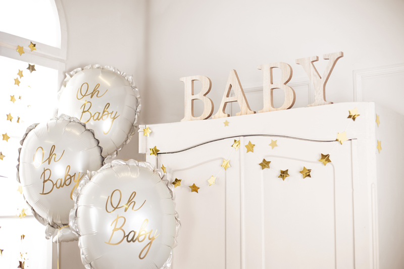 Balões brancos com texto dourado 'Oh Baby' e palavra decorativa 'BABY' em madeira clara sobre móvel branco.