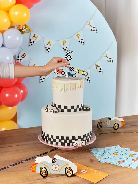 Bolo de aniversário decorado com carros de corrida e padrão xadrez preto e branco, com decoração de festa ao fundo.
