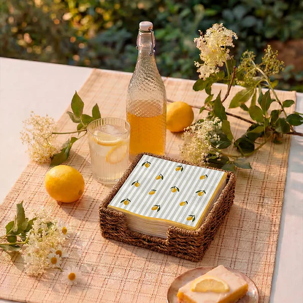 Guardanapos com padrão de limões numa caixa de vime castanho sobre pano bege com limões, flores, garrafa de vidro com líquido amarelo e copo com limonada.