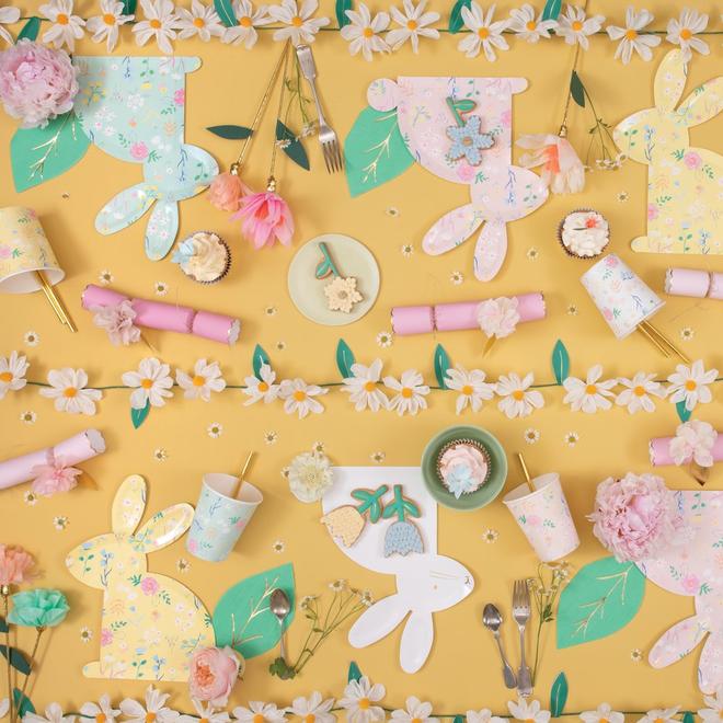 Mesa decorada com coelhinhos de papel, copos, guardanapos e flores sobre fundo amarelo