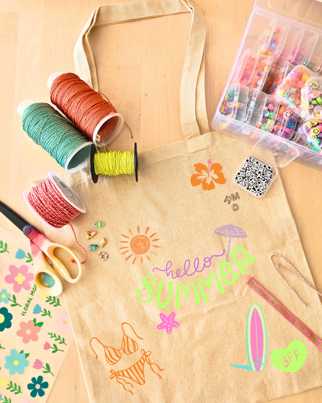 Saco de lona bege decorado com motivos de verão ao lado de rolos de fio, contas coloridas e tesoura sobre mesa de madeira.