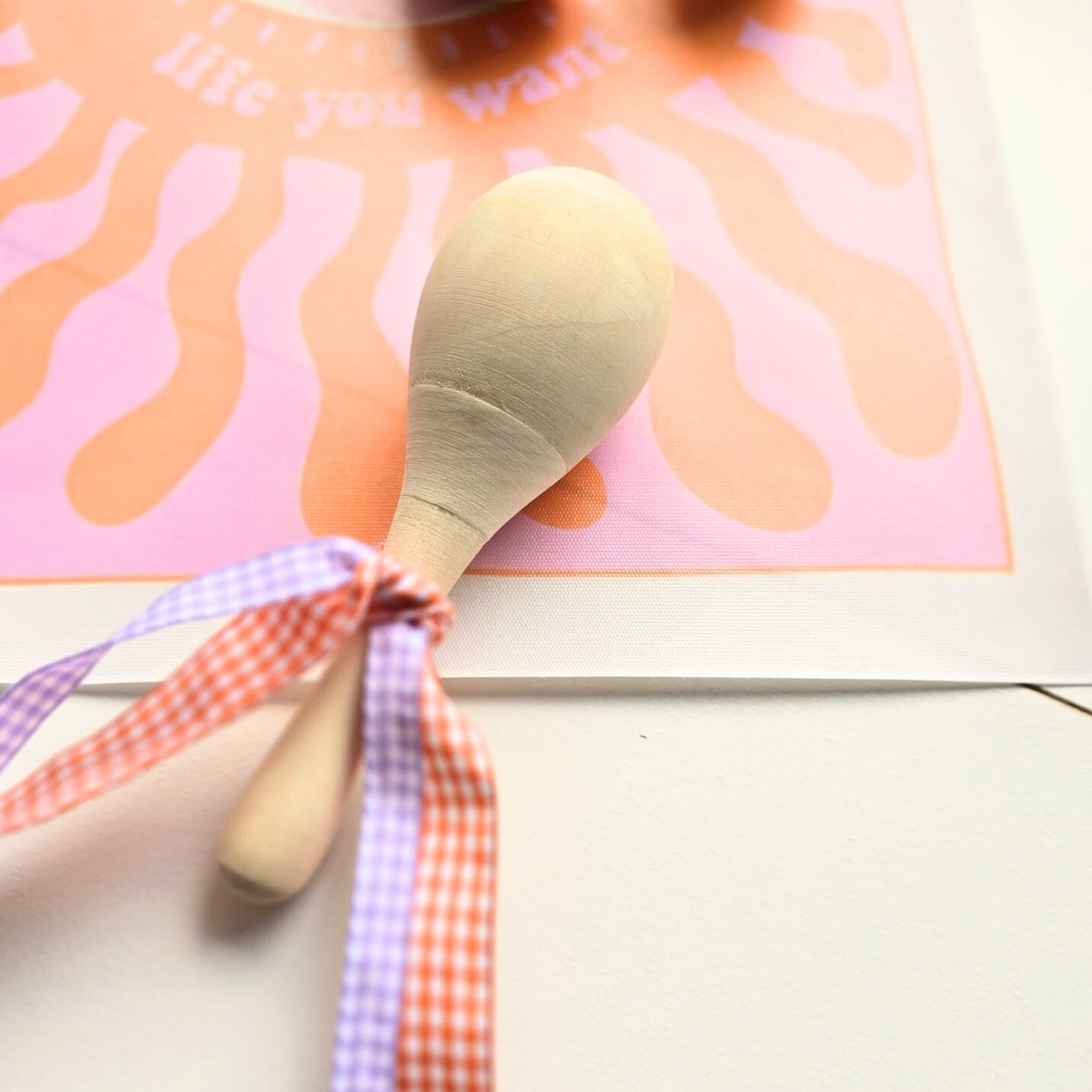 Colher de pau com duas fitas de tecido sobre fundo rosa e laranja com padrão ondulado