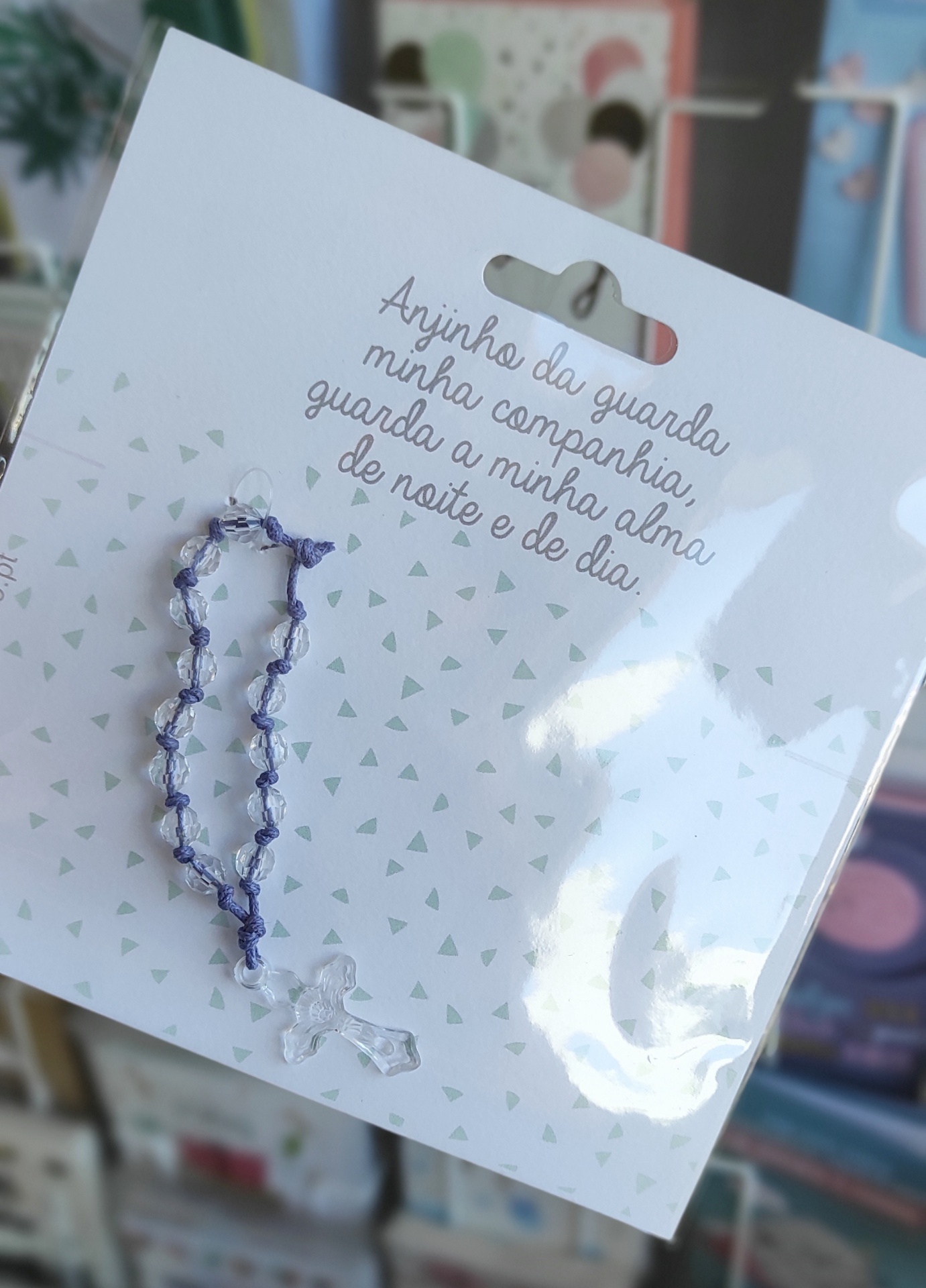 Cartão branco com triângulos cinzentos, pulseira lilás com contas transparentes e crucifixo, com texto em português