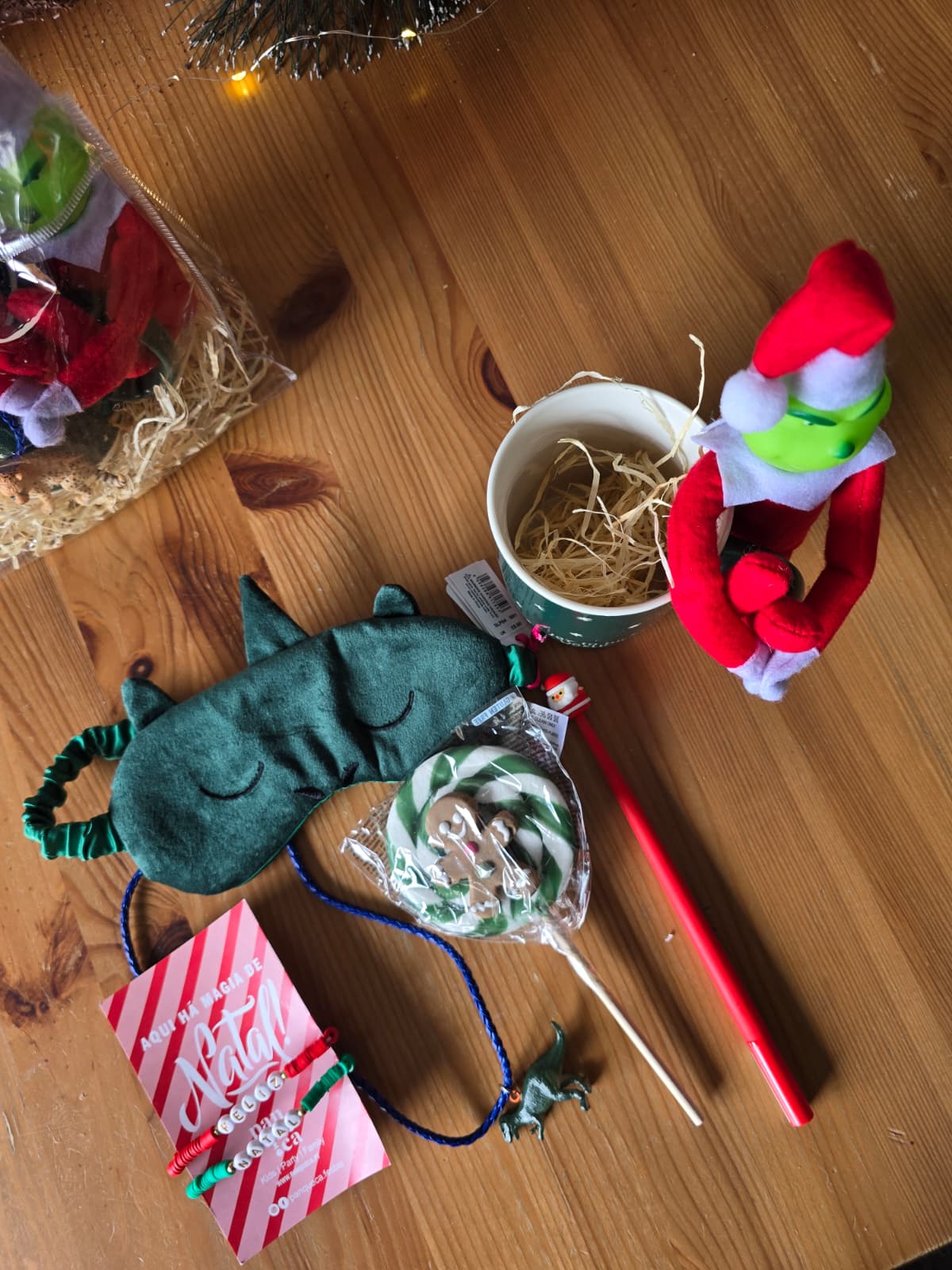Máscara de dormir verde, pirulito, caneta vermelha, pulseira com letras NOEL, boneco de peluche e copo branco sobre mesa de madeira