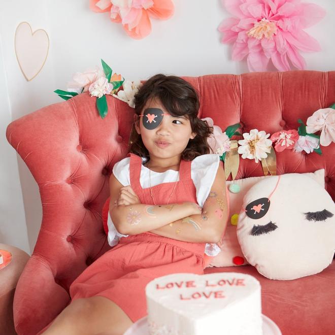 Menina com vestido rosa e tapa-olho preta sentada em sofá rosa de veludo com almofada branca e decoração floral