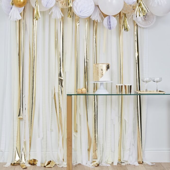 Decoração festiva com balões e fita metálica dourada, mesa de vidro com bolo e copos.