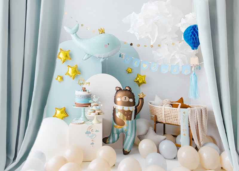 Decoração infantil com cortinas azuis, balões brancos e dourados, baleia azul, nuvens de papel e boneca urso vestida.