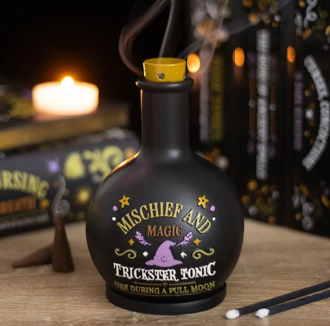 Frasco preto redondo com tampa amarela e rótulo colorido de magia, incenso fumegante e vela acesa num ambiente com livros.
