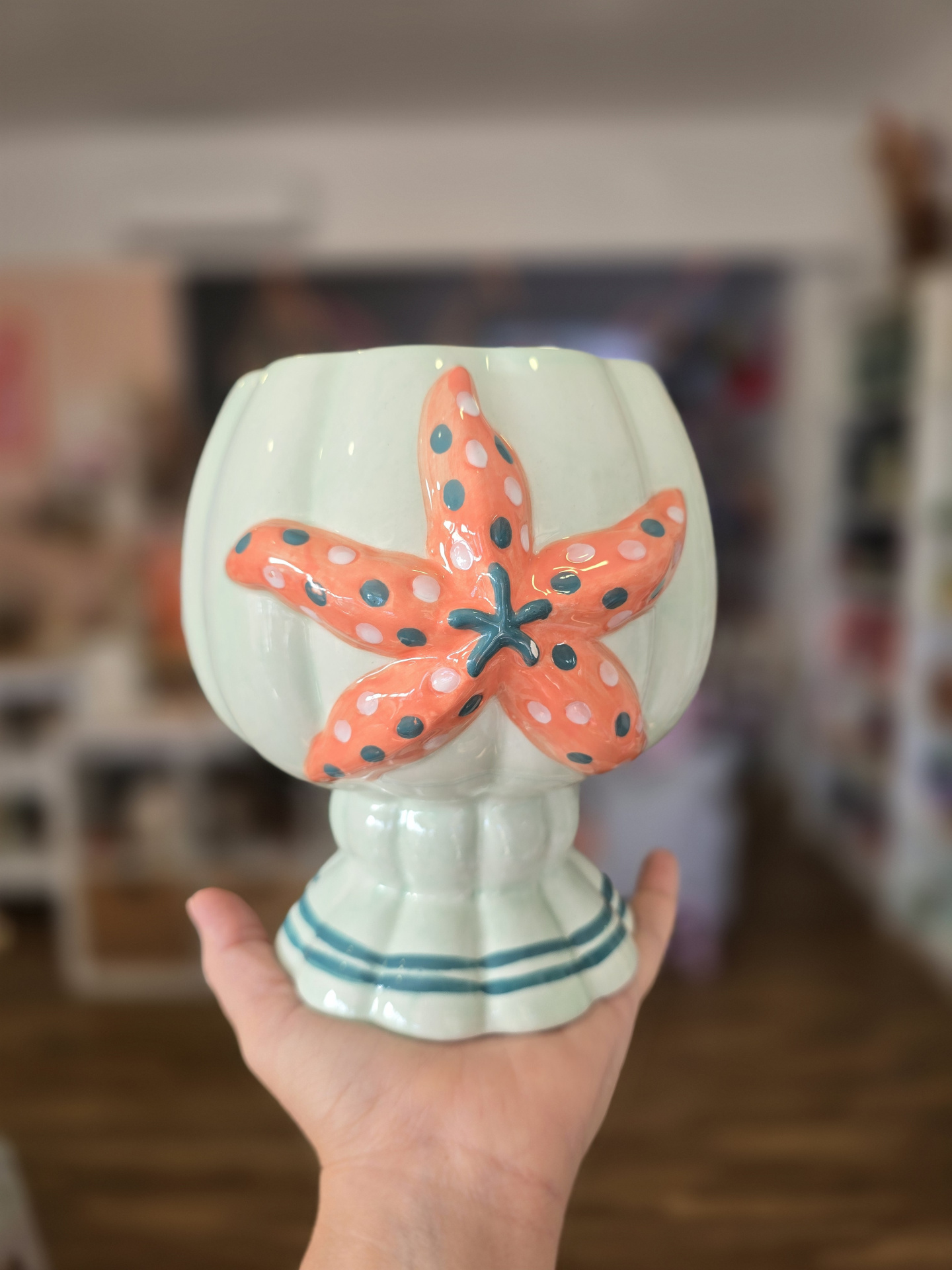 Vaso decorativo branco com estrela do mar em coral e bolinhas, base com riscas azuis, segurado por mão