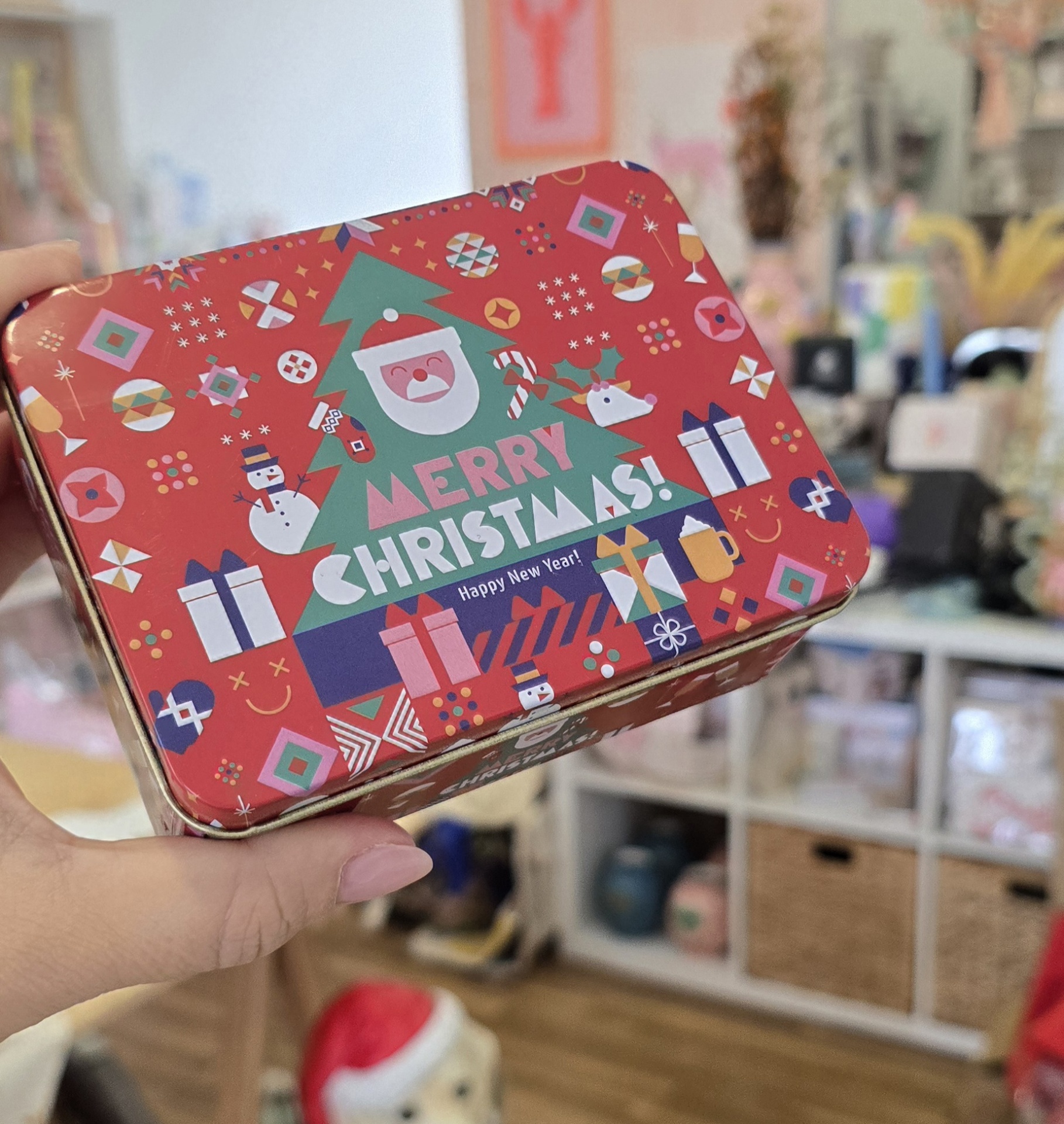 Caixa metálica decorada com motivos natalícios coloridos e texto Merry Christmas segurada por mão