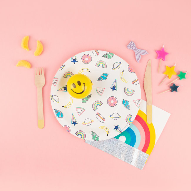 Prato colorido, talheres de madeira, guardanapo com arco-íris e cupcake amarelo sorridente sobre fundo rosa