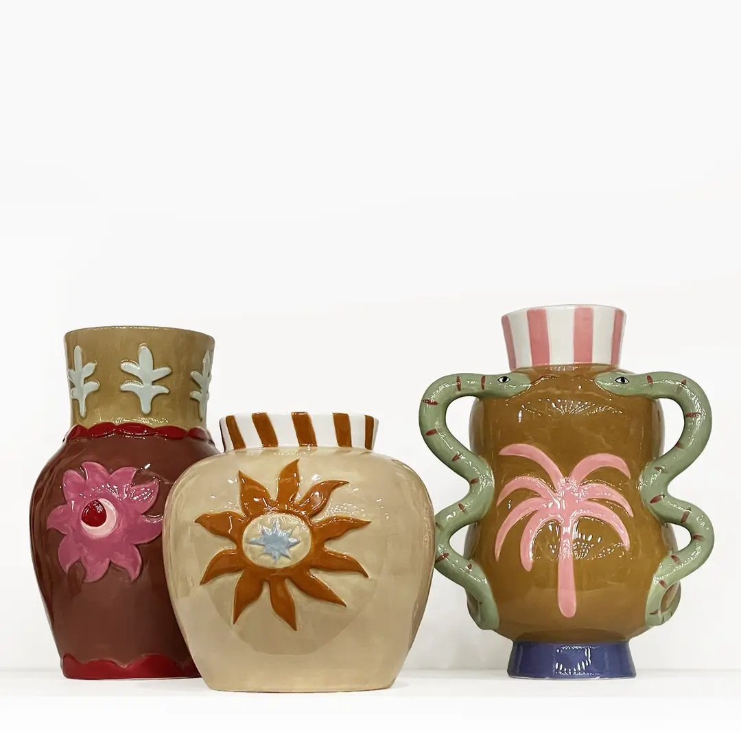Três vasos cerâmicos artísticos com padrões coloridos incluindo flores, sol e palmeira, com alças em forma de serpentes em vaso castanho.