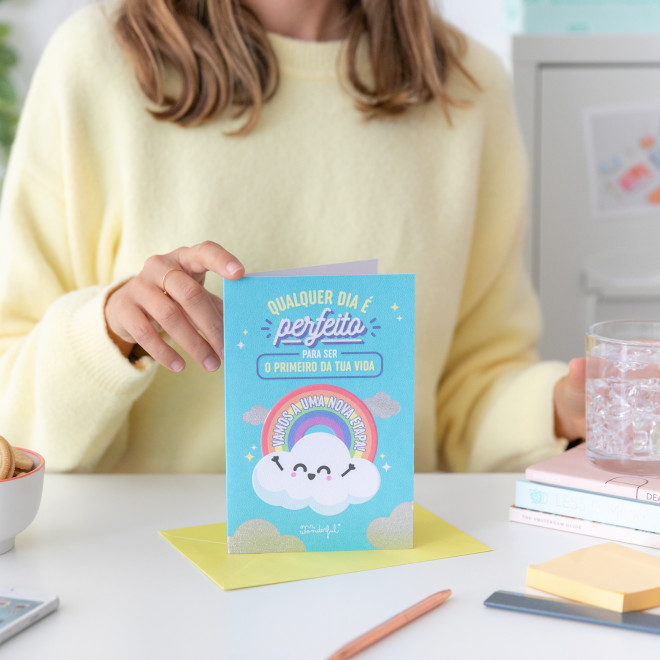 Cartão com arco-íris e nuvem sorridente com texto em português, mãos seguram o cartão sobre mesa clara