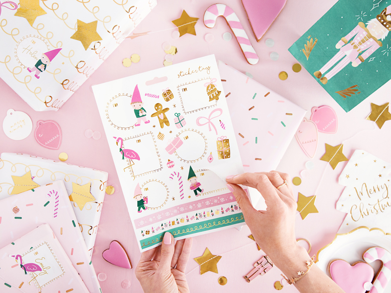 Folha decorativa festiva com desenhos coloridos sendo manuseada sobre mesa rosa com papel de embrulho e confetes