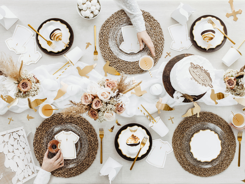 Mesa de celebração com pratos brancos e dourados, bases de mesa em ráfia, bolo branco e flores neutras