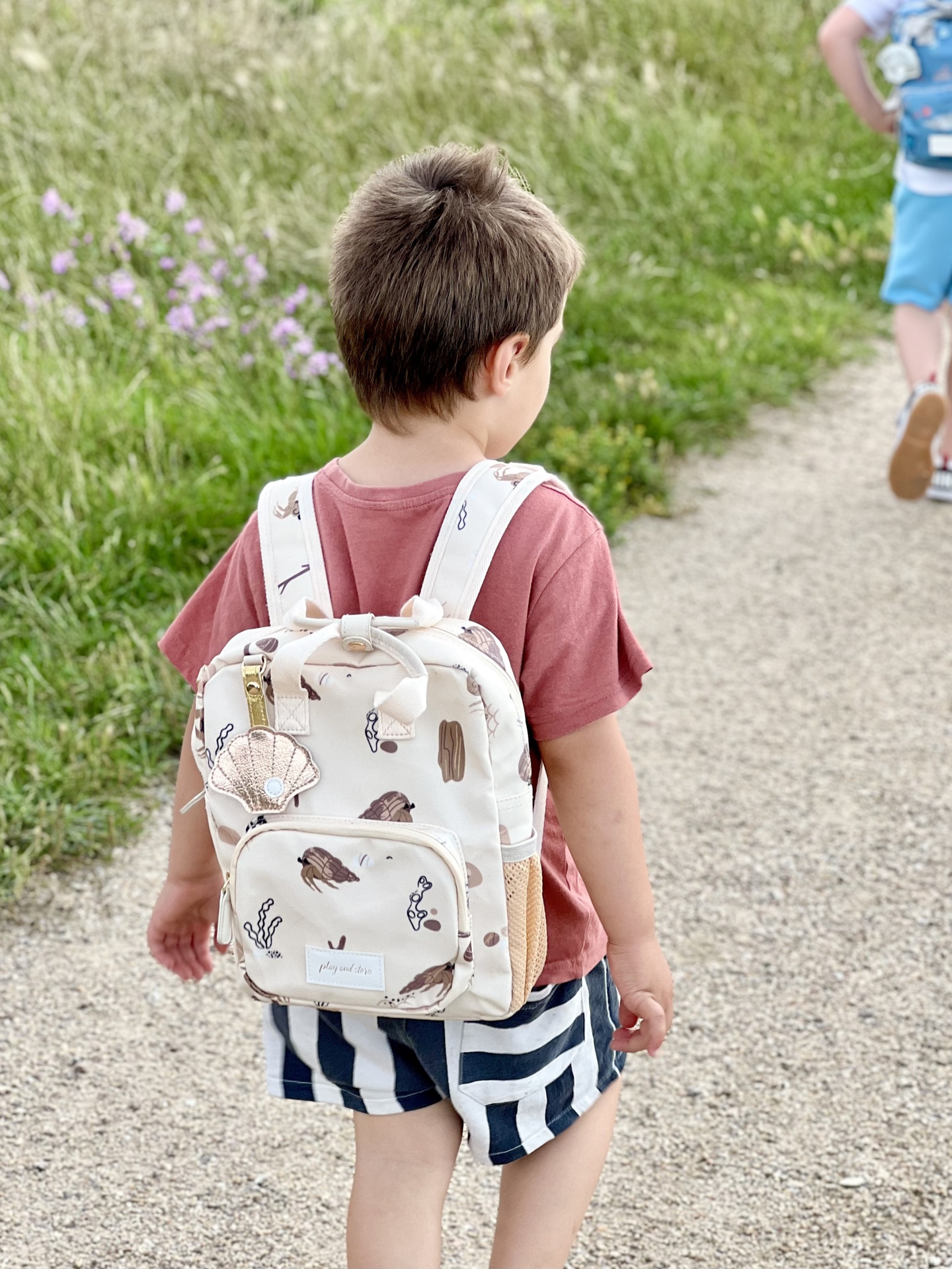 Mochila infantil bege com padrão marinho, usada por criança em trilho ao ar livre