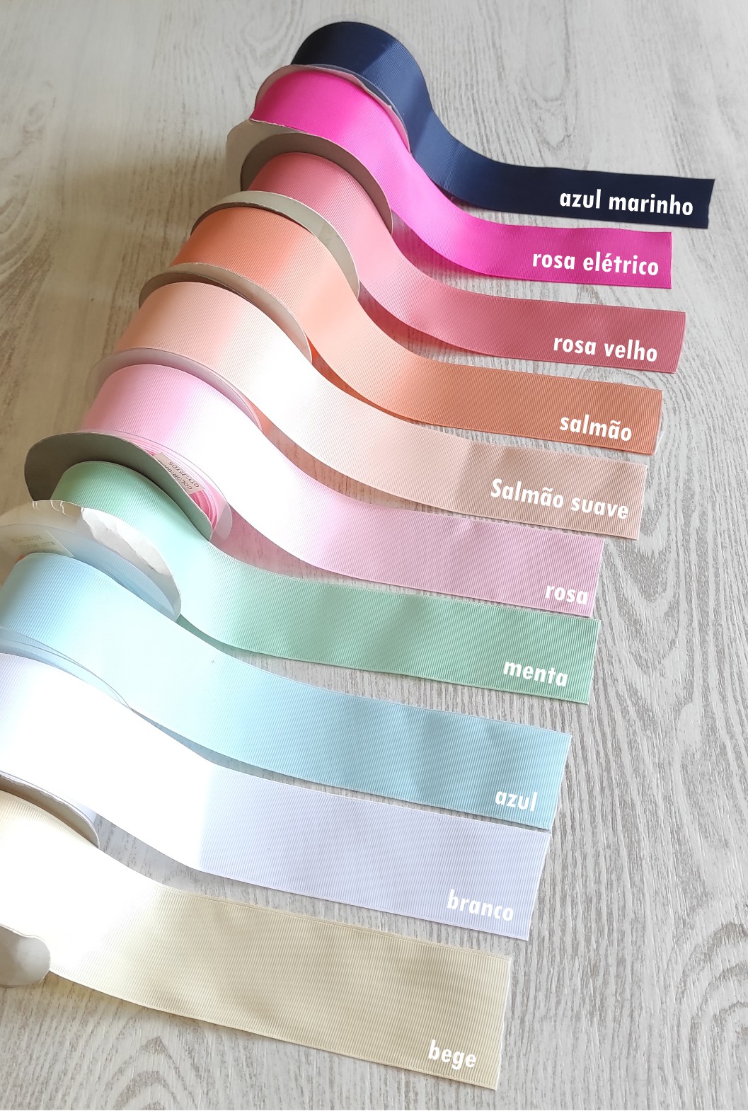 Fitas de tecido coloridas com etiquetas de cores e nomes em português dispostas em superfície de madeira clara.