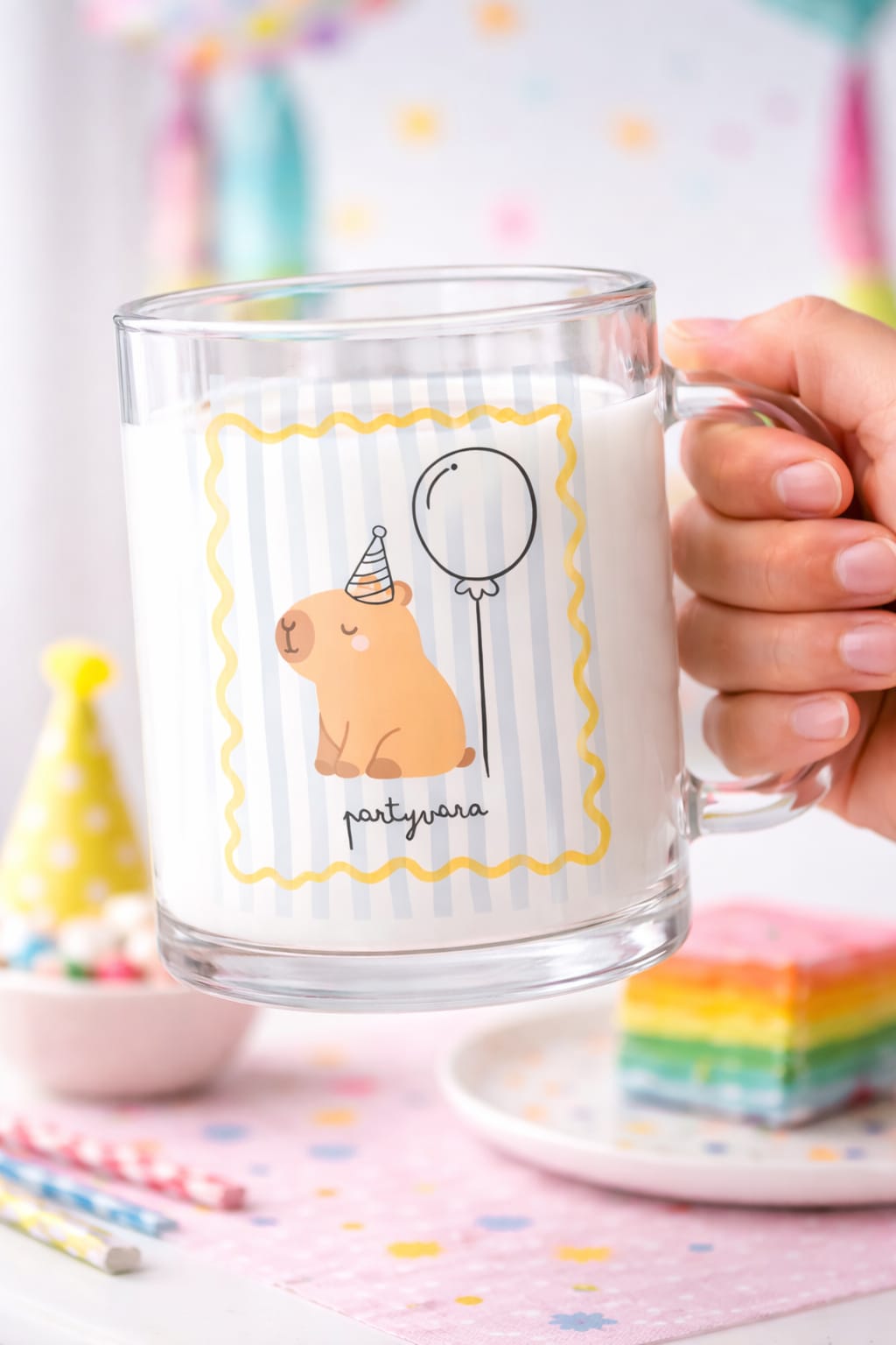Caneca de vidro com desenho de urso de festa e balão, cheia de leite, numa mesa com decoração de festa.