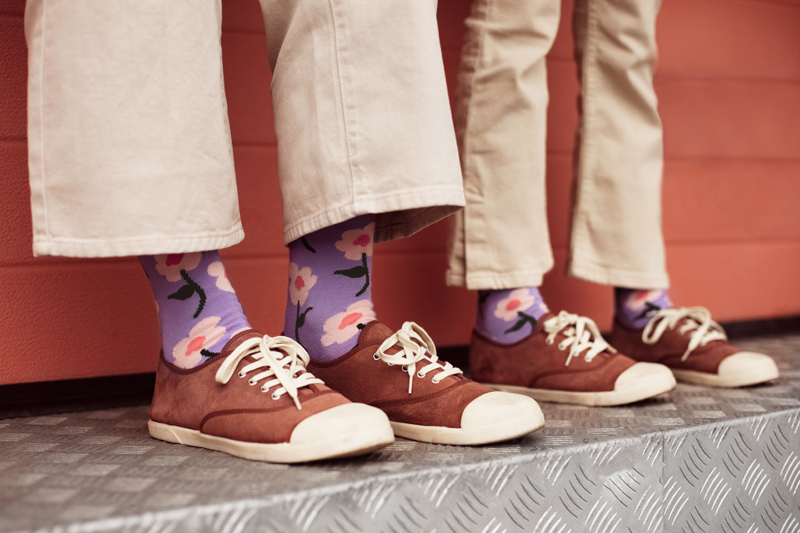 Sapatos castanhos com sola branca, atacadores brancos, meias roxas com flores, calças bege, chão metálico antiderrapante