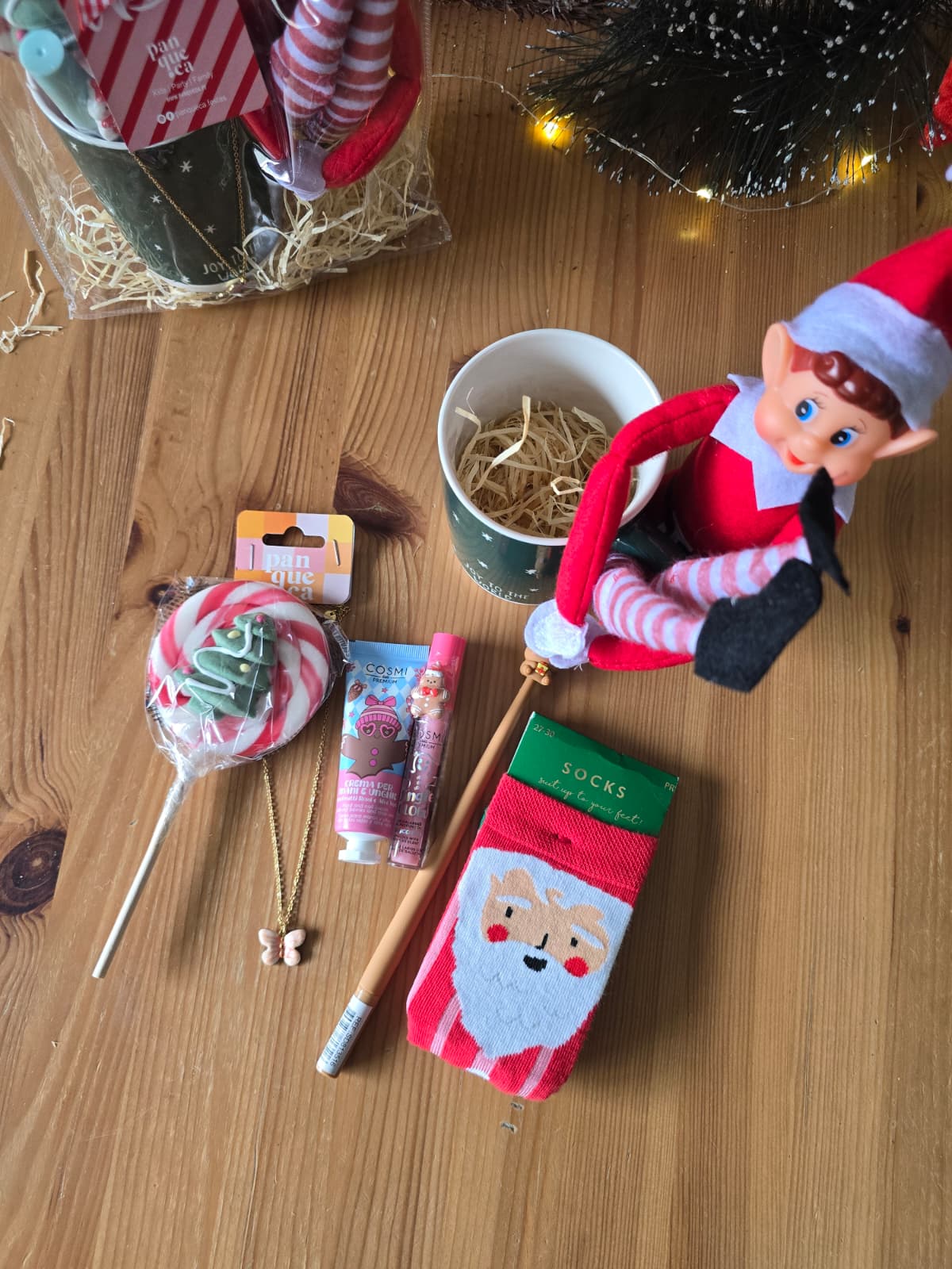 Produtos natalícios variados sobre uma mesa de madeira, incluindo meias, caneca, elfo de tecido, pirulito, creme COSMI, batom, pulseira e lápis.
