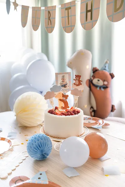 Mesa de festa decorada com bolo branco, balões e enfeites, incluindo um boneco de peluche castanho com chapéu de festa.