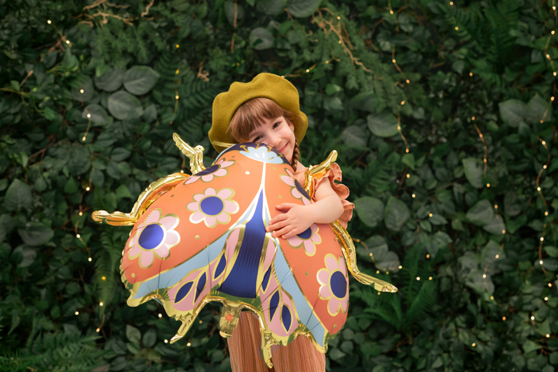 Criança com balão de borboleta colorido e boina mostarda em ambiente com folhagem e luzes