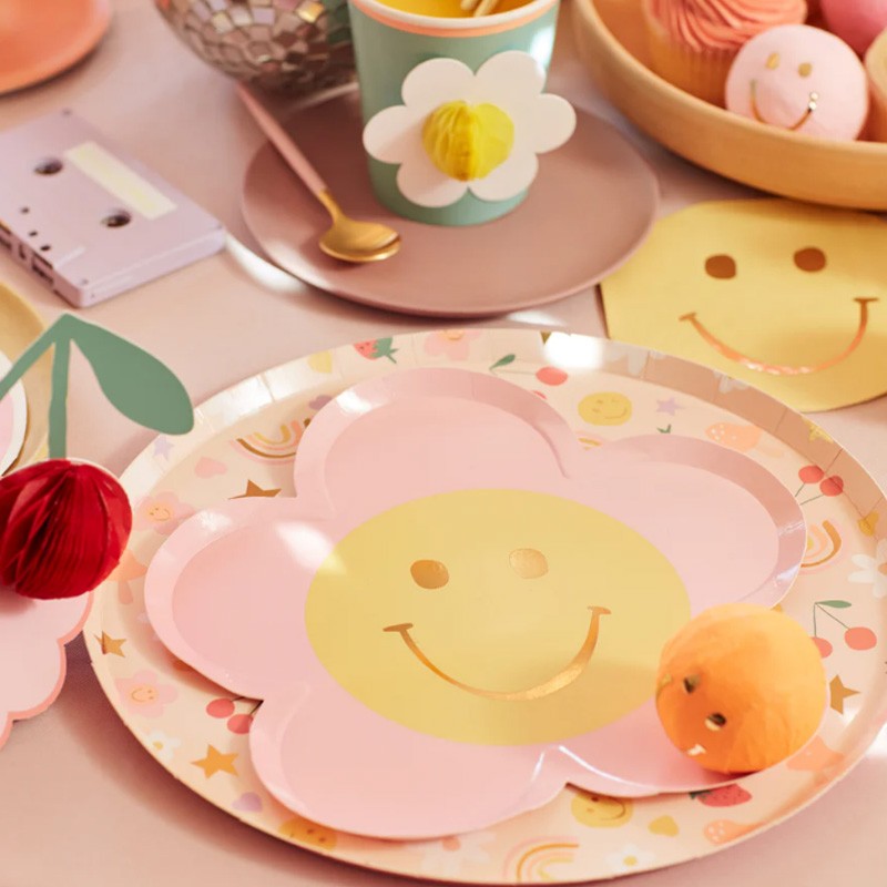 Pratos e copos festivos coloridos com padrões florais e sorriso, guardanapo amarelo sorridente em mesa decorada.