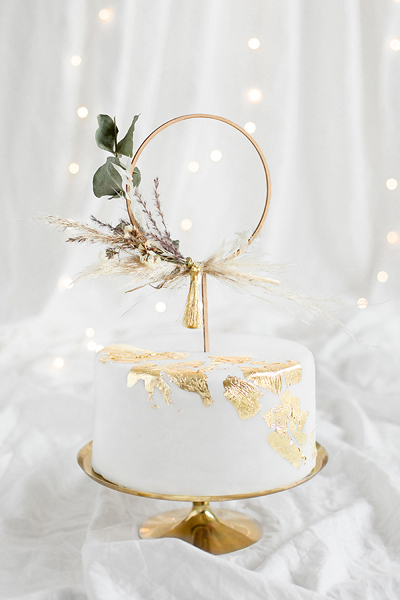 Bolo branco com detalhes dourados e decoração com flores secas sobre suporte dourado