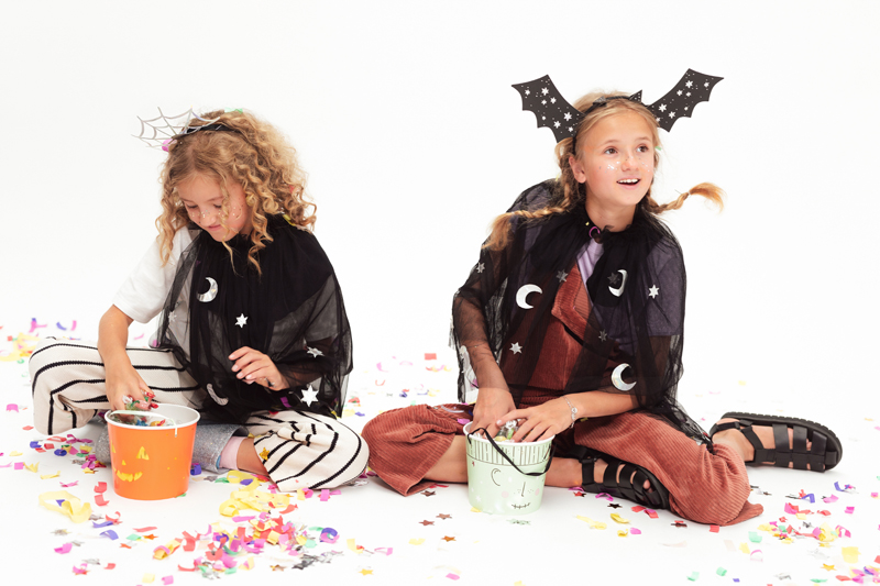 Crianças com capas pretas decoradas com luas e estrelas, sentadas no chão com confetis e segurando baldes coloridos.