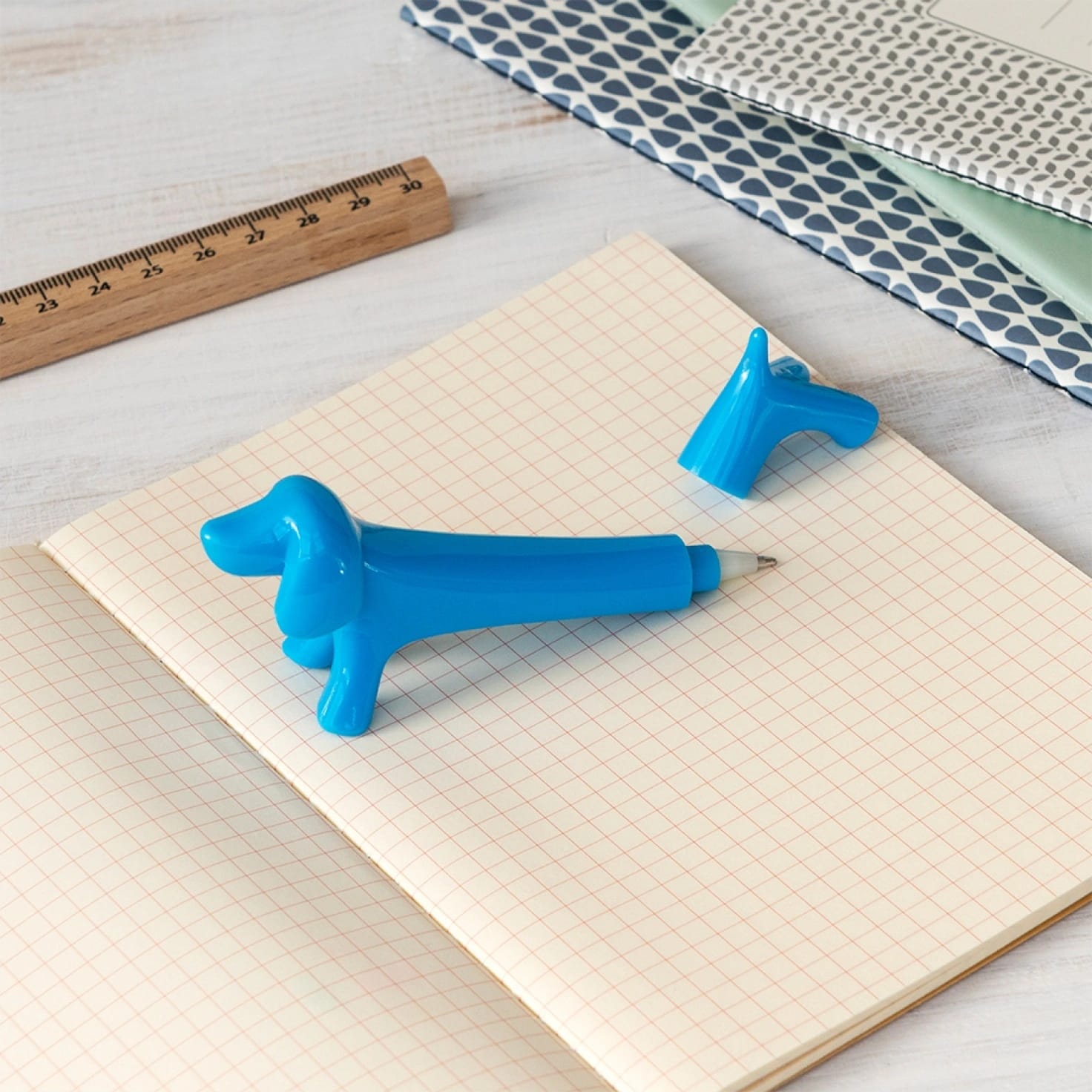Caneta azul com tampa de cão sobre caderno quadriculado e régua de madeira