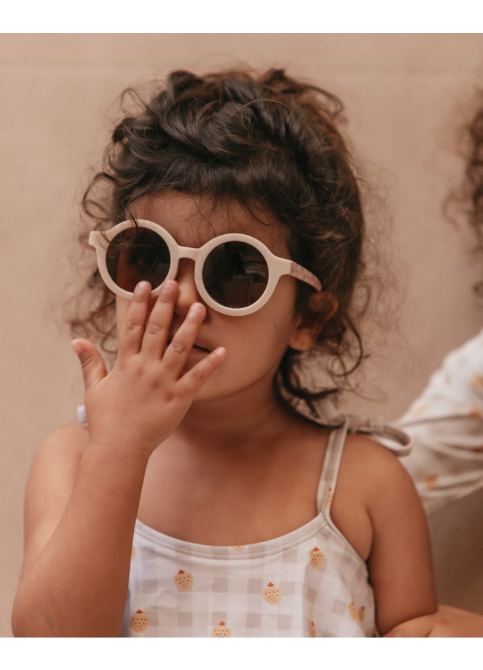 Criança com óculos de sol redondos bege e vestido branco com padrão castanho