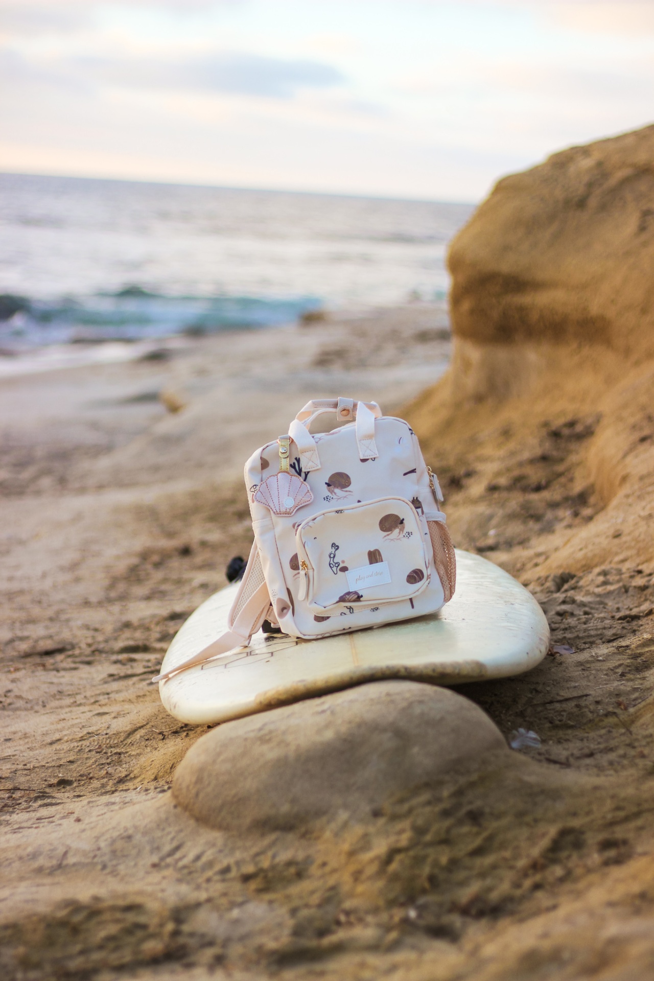 Mochila pequena bege com desenhos de conchas sobre prancha de surf na praia