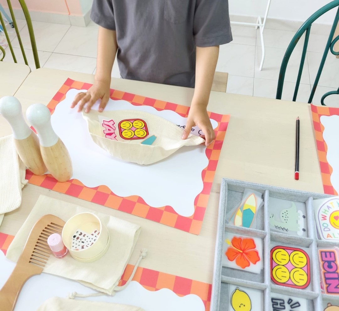 Criança segurando saco de pano com desenho amarelo sorridente e palavra YAY sobre mesa com pentes de madeira, esmalte, adesivos e lápis.